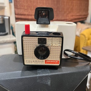 May include: A black and white Polaroid Swinger Model 20 Land Camera with a red button and a black carrying case.