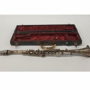 May include: A silver clarinet with intricate detailing, lying horizontally next to its open black case with a red velvet interior. The case has a gold-colored handle. Musical instrument.