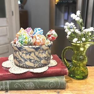 May include: A ceramic bowl with a blue and white marbled pattern filled with assorted Dum Dums lollipops, resting on a white lace doily atop two stacked vintage books, one red and one green, next to a small green glass pitcher with white flowers.