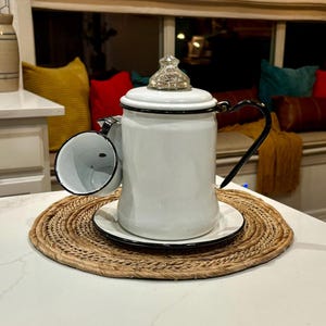 May include: White enamel coffee pot with a black rim and a glass lid. The pot is sitting on a woven placemat with a matching white enamel cup.