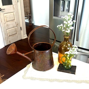 May include: A weathered copper watering can with a curved handle and a perforated spout, positioned next to an amber glass bottle and a small green glass pitcher, both containing sprigs of white baby's breath flowers on a lace-trimmed cloth.
