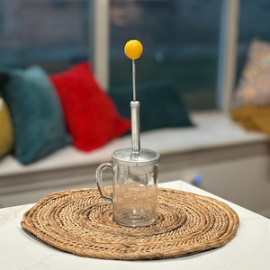 May include: A glass kitchen tool with a yellow knob on top, sitting on a woven round placemat. The tool has a handle and a clear glass container with markings.