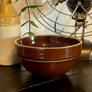 May include: A brown ceramic mixing bowl with a white rim and a brown glaze. The bowl has a slightly rounded shape and a wide, flat base.