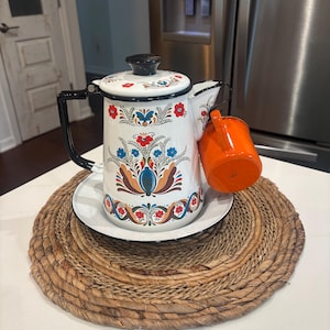 May include: White enamel coffee pot with black handle and lid, decorated with red, blue, and orange floral patterns. An orange mug rests on the side. The pot sits on a white plate and a woven placemat.