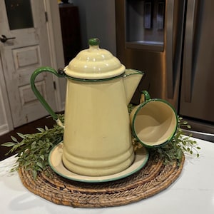 May include: A pale yellow enamel coffee pot with a matching saucer and a single cup. The pot features a domed lid, a green handle, and green trim along the edges. The cup is suspended from the spout of the pot, also featuring green trim.