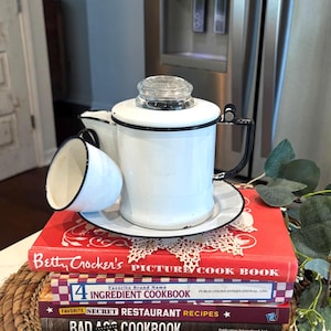 May include: A white enamel coffee pot with a glass lid and a matching cup, both with black trim, sit on a saucer. The pot and cup are on top of a stack of cookbooks, including "Betty Crocker's Picture Cook Book."