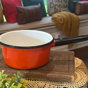 May include: A small, orange enamel saucepan with a black handle and rim. The interior is white. The pan sits on a vintage book and a decorative doily. The background includes a bench with pillows and a table.