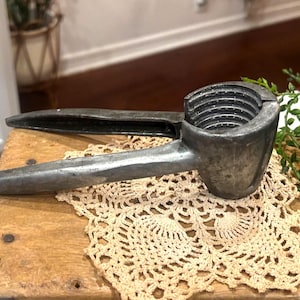 May include: Vintage metal nutcracker with a textured, silver finish. The nutcracker is resting on a wooden surface with a beige lace doily. Greenery is to the right of the nutcracker.