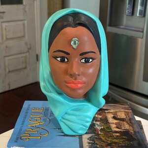 May include: A ceramic bust of a woman with dark skin, wearing a turquoise head covering. The bust has a jewel on the forehead and is resting on a stack of books. The books have the word "Prague" on them.