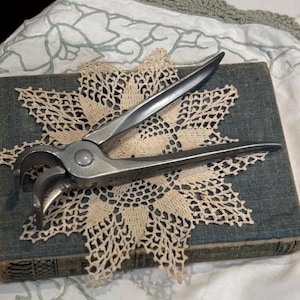 May include: A pair of silver-toned metal end-cutting pliers with a pivot point and curved jaws. The tool features long, tapered handles and a metallic finish, positioned centrally on a rectangular blue book and a beige crocheted lace doily.
