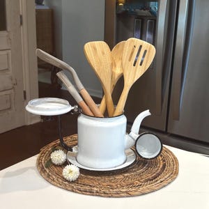 May include: A white enamel teapot with a black rim and handle, functioning as a utensil holder. It contains several bamboo kitchen tools, including spatulas and a basting brush, alongside a grey silicone spatula. Two small white flowers sit nearby.