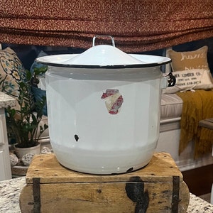 May include: A white enamelware pot with a lid, black trim, and handles. The pot has a rounded shape and sits on a wooden box. A faded sticker is on the front of the pot.