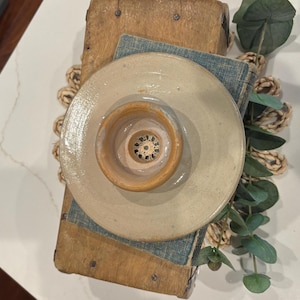 May include: A beige ceramic jar lid with a central, smaller, tan-colored bowl. The lid sits atop a small, blue-covered book and a wooden box. Decorative elements include a woven mat and eucalyptus sprigs.