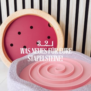 Könnte beinhalten: Zwei rosa und beigefarbene Stapelspielzeug-Basen mit einem Spiraldesign. Der Text "3...2...1 WAS NEUES FÜR EUERE STAPELSTEINE!" befindet sich auf der Basis.