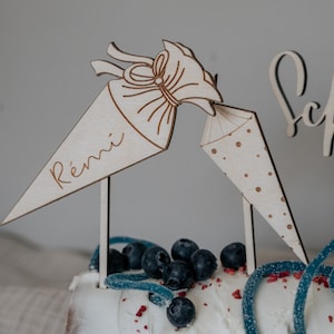 Könnte beinhalten: Holz-Kuchentopper mit zwei Kegeln, einer mit dem Namen "Rémi" und der andere mit Punkten. Die Kegel stehen auf einem Kuchen, der mit Blaubeeren und blauen Süßigkeiten verziert ist.