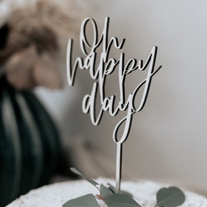 Könnte beinhalten: Ein weiß-schwarzer Tortenaufsatz mit den Worten "Oh happy day" in Schreibschrift. Der Aufsatz befindet sich auf einem Kuchen mit Puderzucker und ist mit grünen Blättern verziert.