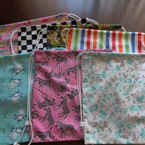 May include: A collection of drawstring bags with various patterns. Designs include pink with zebras, teal with butterflies, and a black and white checkered pattern. Each bag has a white drawstring closure.