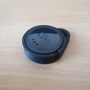 May include: A black Tile Bluetooth tracker with the word "Tile" printed on the front. The tracker has a small hole on the side for attaching to a keychain.