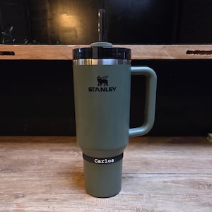 Peut inclure: Un mug Stanley vert avec un couvercle et une poignée noirs. Le mug porte un bandeau noir avec le nom "Carlos". Le logo Stanley est visible à l'avant. Une paille transparente est insérée dans le couvercle.