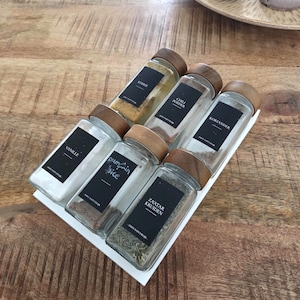 Könnte beinhalten: Ein Set aus sechs rechteckigen Gewürzgläsern aus Glas mit Holzdeckeln, angeordnet auf einem weißen Tablett. Jedes Glas hat ein schwarzes rechteckiges Etikett mit weißem Text, der Kerrie, Chili Poeder, Koriander, Vanille, Pumpkin Spice und Za'atar Kruiden lautet.
