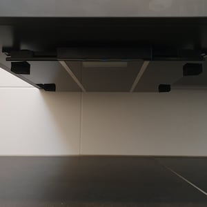 Può includere: Sotto un mobile nero con un ripiano in vetro e staffe nere. Il mobile è montato su una parete piastrellata bianca.