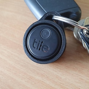 Op de afbeelding: Een zwarte Tile Bluetooth-tracker met een witte knop en het woord "tile" erop gedrukt. De tracker is bevestigd aan een zilveren sleutelhanger.