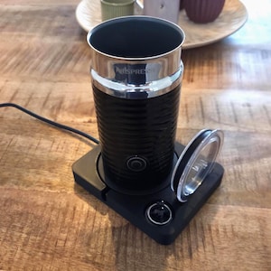 Peut inclure: Un mousseur à lait Nespresso noir avec un corps cylindrique, un bord supérieur en métal argenté et un bouton d'alimentation circulaire sur le devant. L'appareil repose sur une base carrée noire avec un cordon et un couvercle transparent.