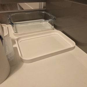Könnte beinhalten: Ein weißes rechteckiges Tablett mit abgerundeten Ecken und einem erhöhten Rand. Ein klarer rechteckiger Glasbehälter steht auf einem weißen Tablett darüber. Die Artikel befinden sich auf einer weißen Oberfläche.