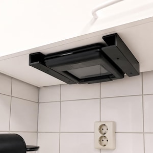 May include: A black under-cabinet mount bracket securely holding a square black device. The mount features two parallel side supports attached to the underside of a white cabinet, providing a streamlined storage solution for the electronic unit.