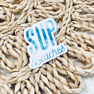 May include: A white sticker with the words "SUP Beaches" in blue, with a watercolor effect. The sticker is on a woven, light brown surface. The text is in a stylized font, suggesting a beach or water activity theme.