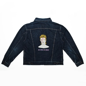 Peut inclure: Une veste en jean bleu foncé avec une image brodée de David Bowie au dos. L'image comprend le texte "We Could Be Heroes".