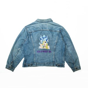 Peut inclure: Une veste en jean bleue avec une image brodée d'une femme portant une couronne bleue et blanche et tenant un téléphone. Le texte "ALLO ST-HUBERT BBQ" est brodé sous l'image.