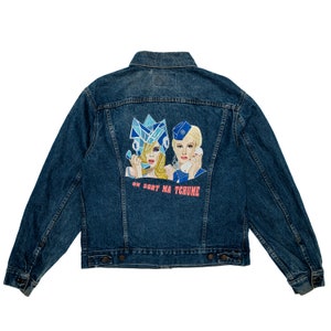 Può includere: Una giacca di jeans blu con una toppa ricamata sul retro. La toppa presenta due donne con capelli e vestiti colorati, e il testo "ON SORT MA TCHUME".