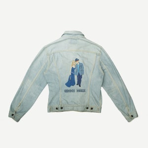 Può includere: Giacca di jeans blu chiaro con un'immagine ricamata di una coppia in abito da sera. Il testo "Gimme More" è ricamato sotto l'immagine.