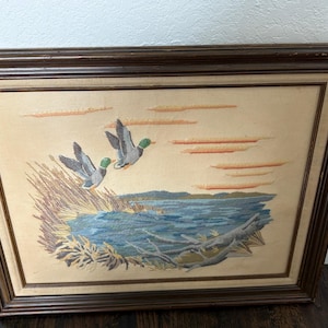 May include: Embroidered artwork of two ducks in flight over a lake scene. The ducks have gray and white wings and green heads. The artwork is framed in a dark brown wooden frame, with a beige background and blue water.