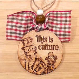 May include: Wooden ornament with the phrase "This is culture" and a cartoon illustration. The ornament has a red and white checkered ribbon bow and a twine loop for hanging. The ornament is round.