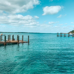 Puede incluir: Un muelle de madera se extiende hacia un cuerpo de agua turquesa tranquilo. El cielo es de un azul brillante con nubes blancas esponjosas.