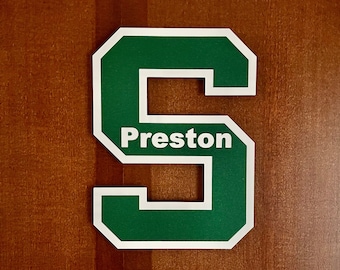 May include: A green block letter S with a white border and the name Preston printed in white across the center. The letter features a bold, athletic-style font and is mounted on a flat surface, creating a clear and legible display of the name.