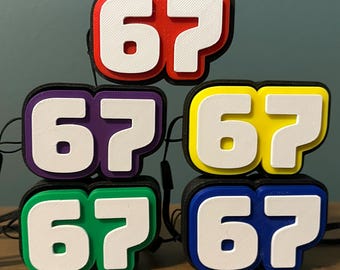 May include: A stack of colorful, numbered accessories, each displaying the number "67" in white against a vibrant background. The accessories are in red, purple, yellow, green, and blue, with black borders. A black lanyard is attached to each.