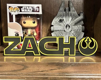 May include: A black and yellow nameplate with the name "ZACH" on it. The nameplate is decorated with a Star Wars Rebel Alliance symbol and a model of the Millennium Falcon.