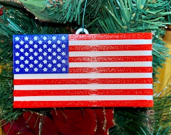 May include: A red, white, and blue American flag ornament with a glittery texture. The flag is hanging from a Christmas tree.