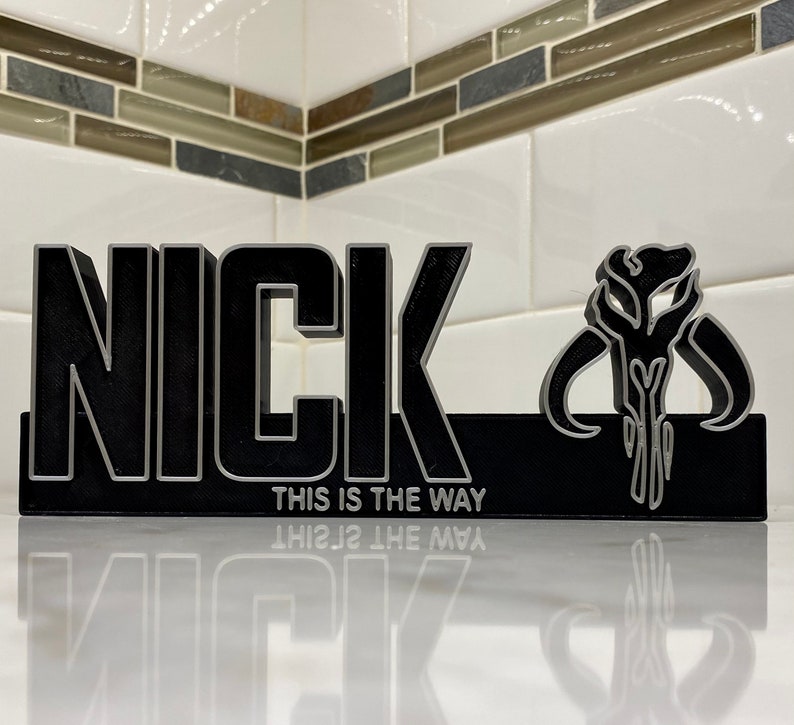 May include: A black and silver 3D printed nameplate with the name "NICK" and the Mandalorian symbol. The text "This is the way" is printed below the name.