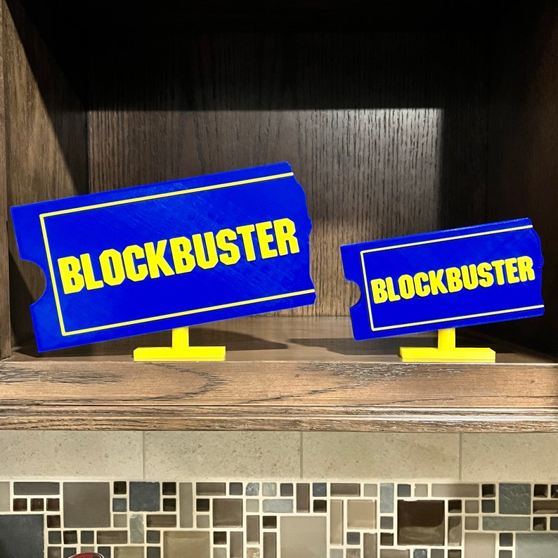 May include: Two blue and yellow Blockbuster signs on a wooden shelf. The signs are shaped like movie ticket stubs.