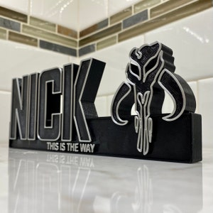 May include: A black 3D printed nameplate with the name "NICK" in silver lettering. The nameplate also features a silver Mandalorian symbol and the text "THIS IS THE WAY" in silver lettering.