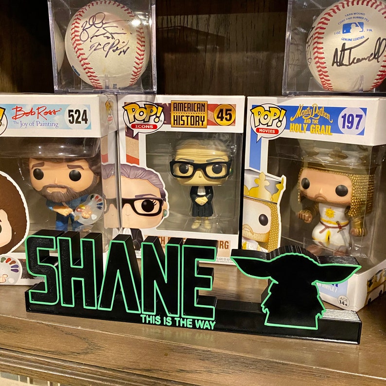 May include: A black and green neon sign with the word "SHANE" and the text "THIS IS THE WAY" below. The sign is sitting on a shelf with several Funko Pop! figures in front of it.