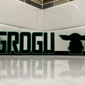 May include: Black and green wooden sign with the word "GROGU" in large letters and a silhouette of the character Grogu from the Star Wars series The Mandalorian.