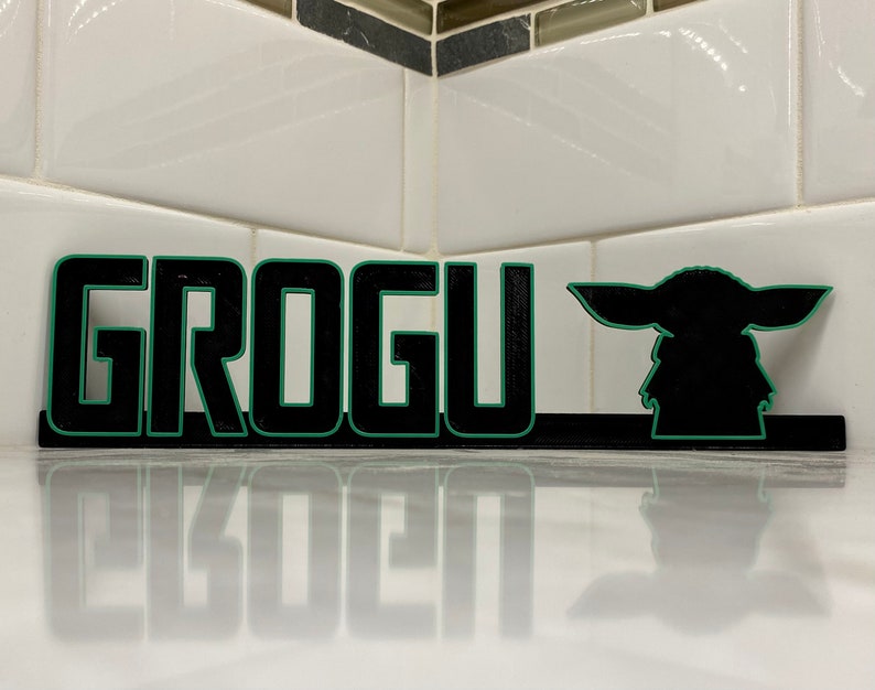 May include: Black and green wooden sign with the word "GROGU" in large letters and a silhouette of the character Grogu from the Star Wars series The Mandalorian.