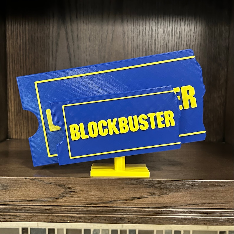 May include: A blue and yellow 3D printed sign that reads "BLOCKBUSTER" on a wooden shelf.