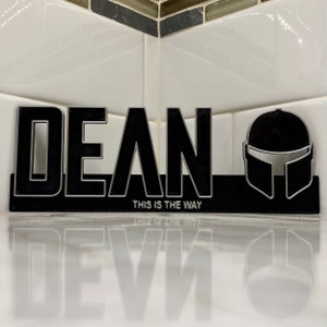 May include: A black and silver 3D printed sign with the name "DEAN" and the phrase "This is the Way" underneath. A Mandalorian helmet is on the right side of the sign.