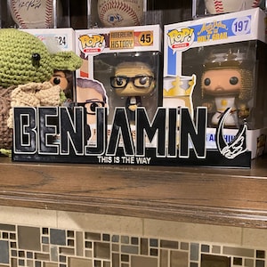 May include: A black and white 3D printed sign with the name "BENJAMIN" in bold letters and the phrase "THIS IS THE WAY" underneath. The sign is sitting on a shelf with a variety of pop culture figurines behind it.